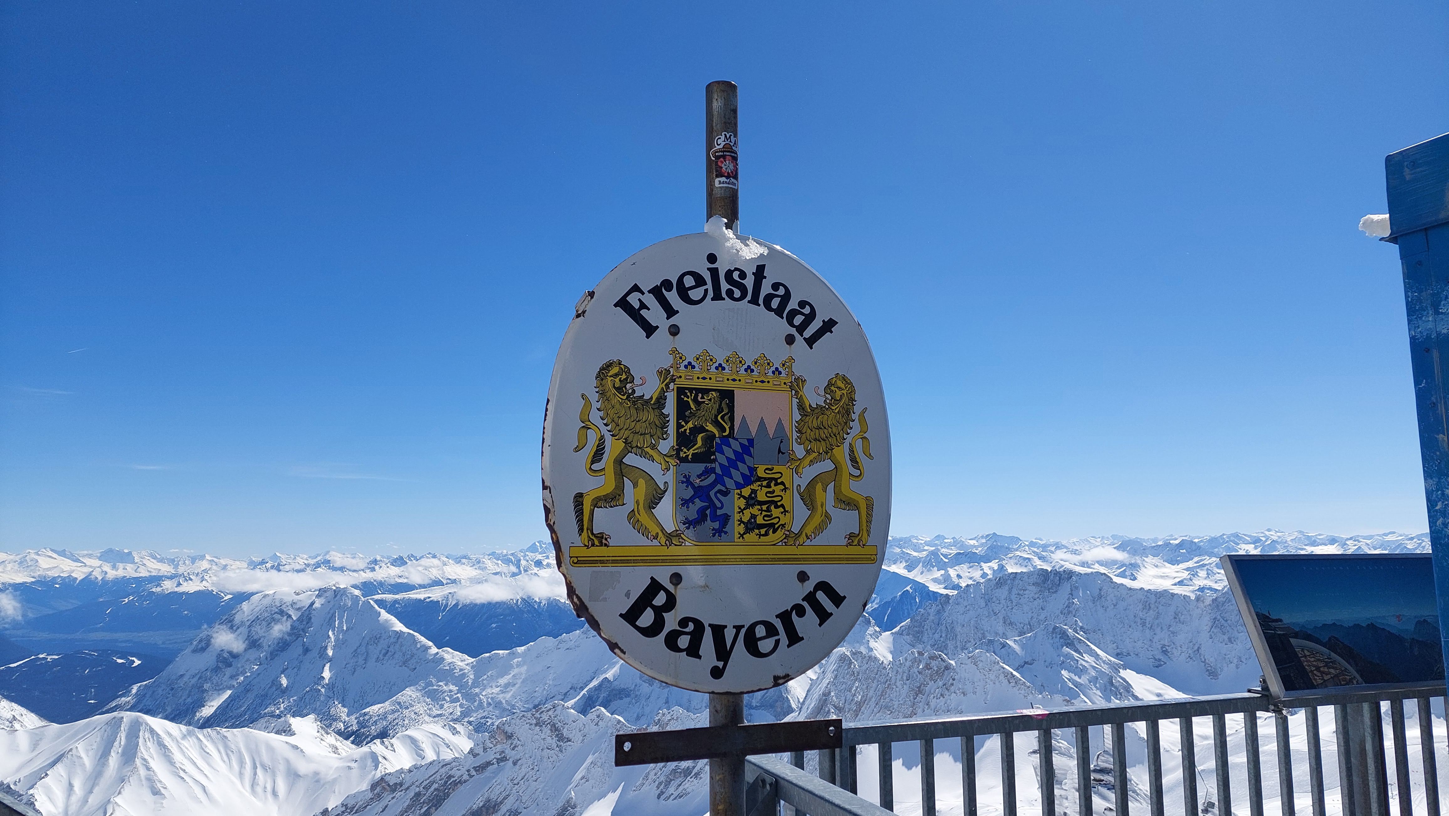 An oval sign reading 'Freistaat Bayern' mounted on a railing at the Zugspitze summit, with a panorama of snow-covered mountains behind