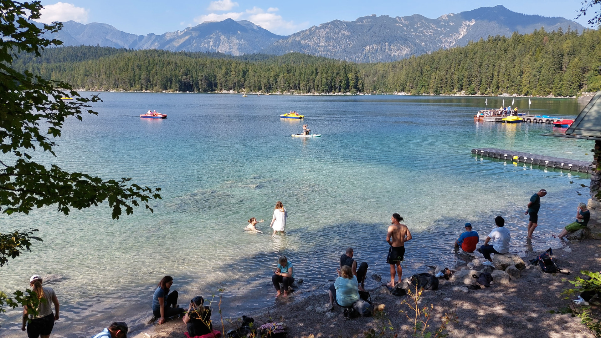 People wading and swimming in the turquoise water of Eibsee lake on a sunny September day, with colorful pedal boats and the Wetterstein mountains behind