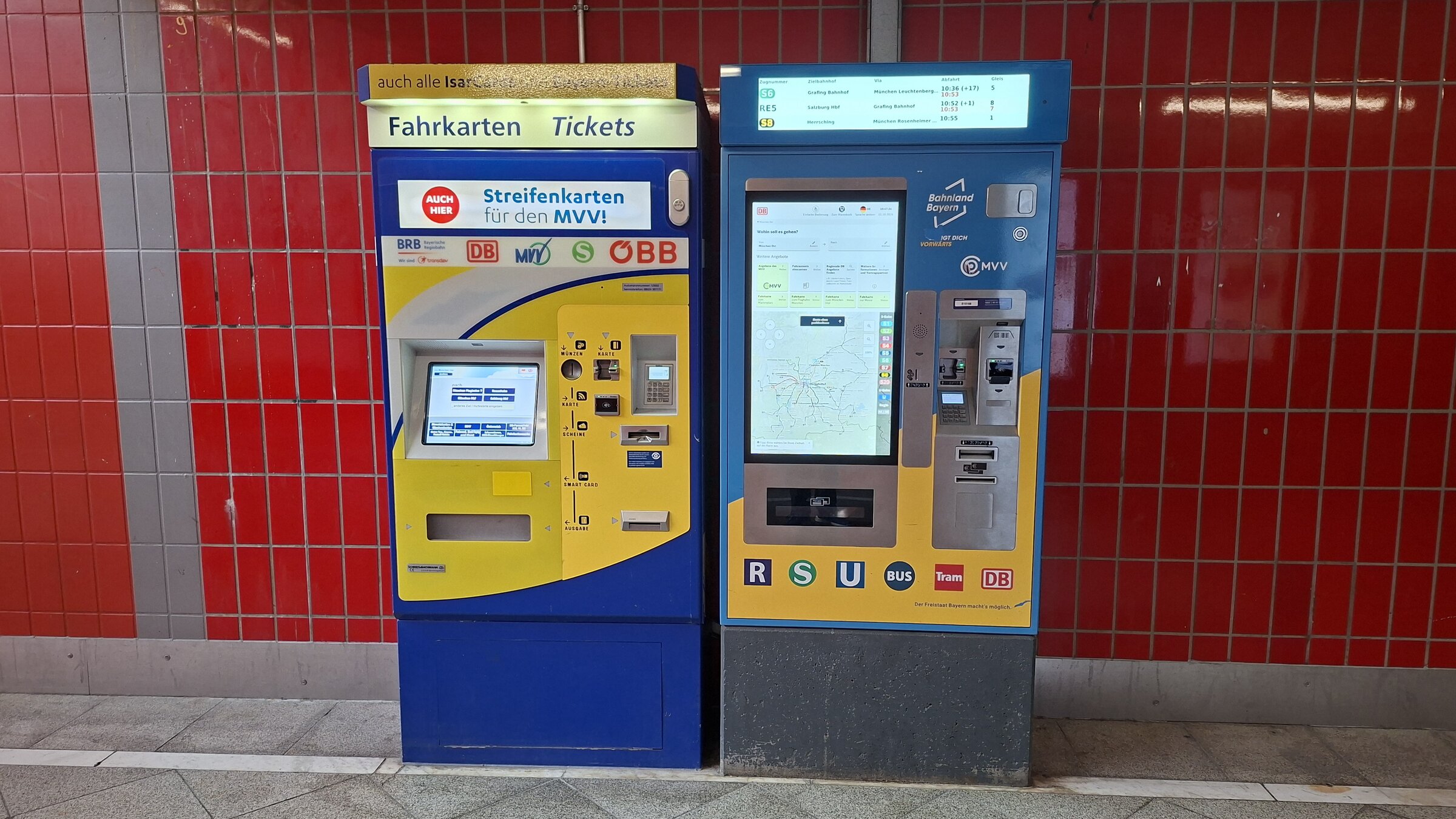 A DB ticket machine and an MVV ticket machine side by side at a Munich train station, with a departure board showing regional trains overhead