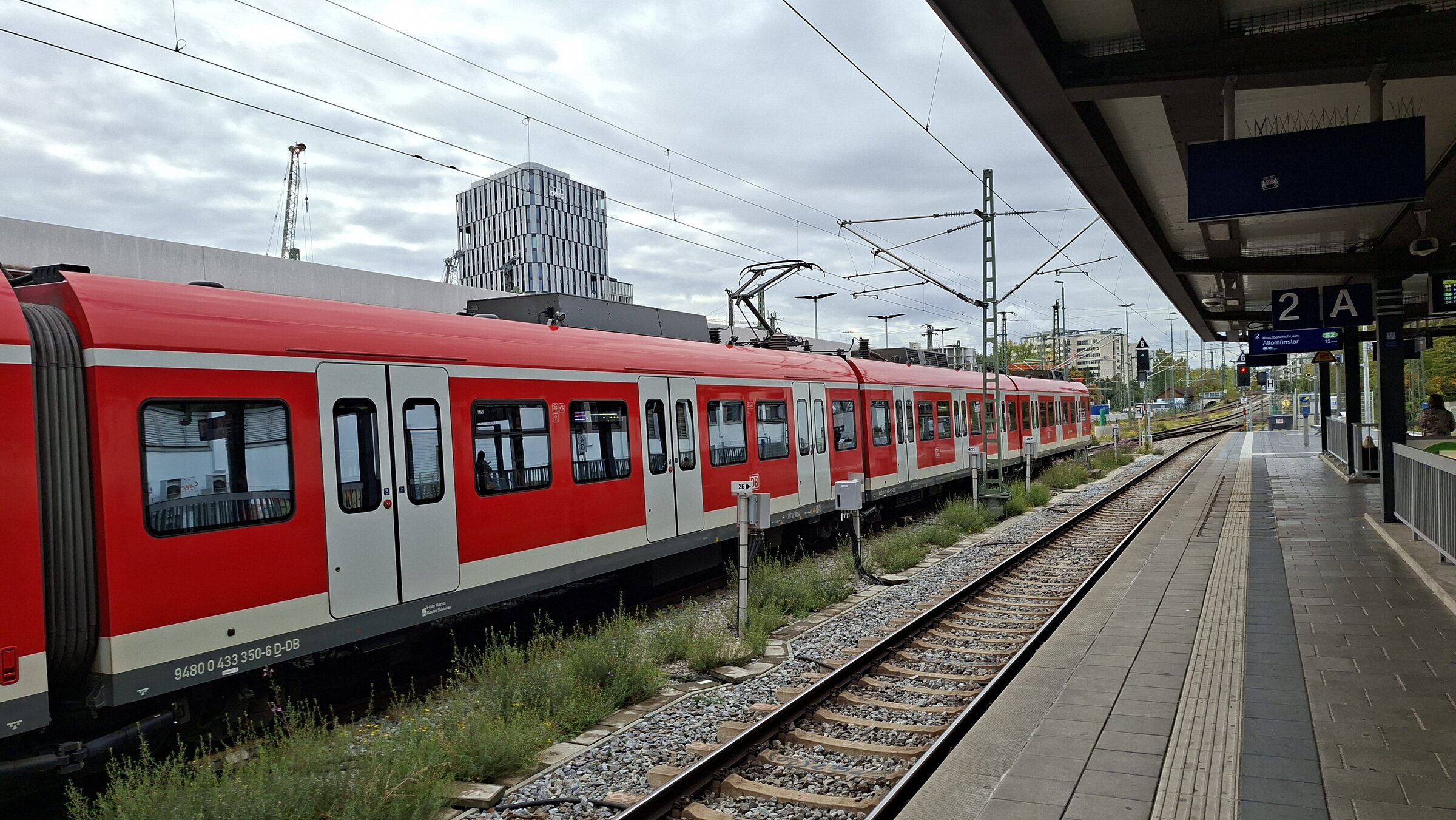 An S-Bahn train waiting at a Munich platform, with the city skyline and construction cranes in the background