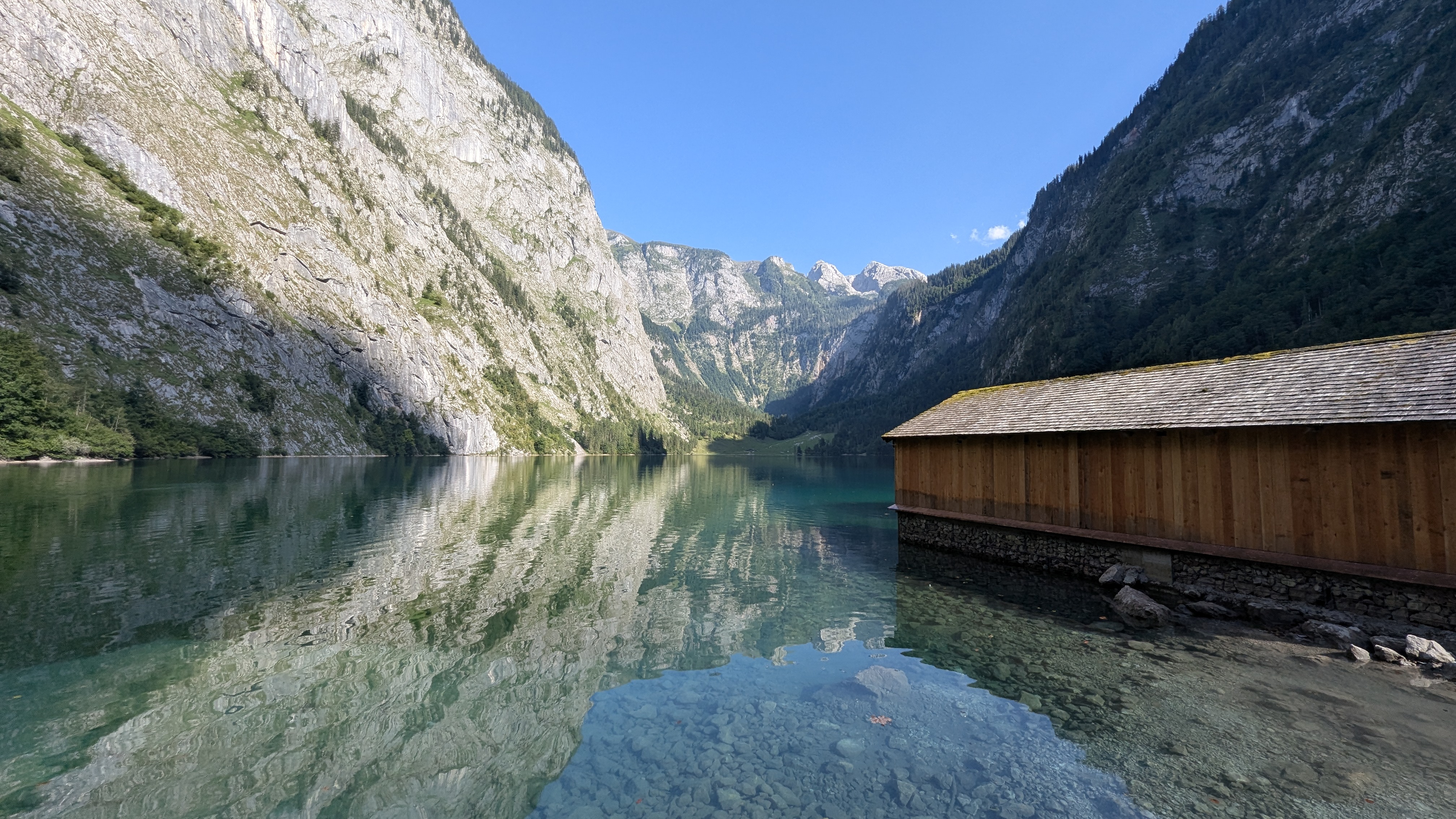 Obersee lake near Königssee, with steep Alpine cliffs reflected in clear water and a wooden boathouse on the shore.