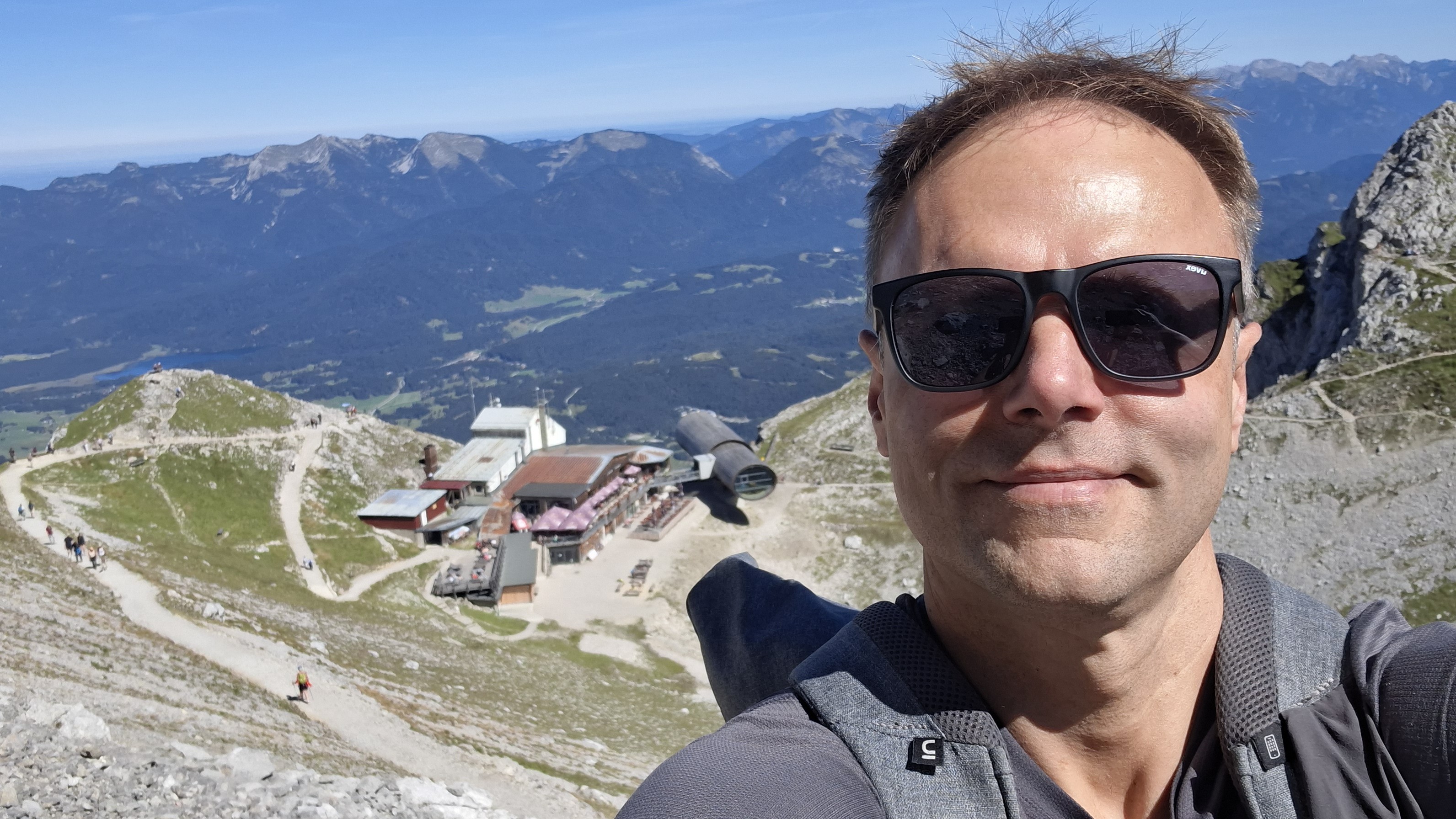 Martinova selfie na vrchole so slnečnými okuliarmi a sivými popruhmi ruksaku, v pozadí horná stanica lanovky Karwendelbahn a alpské údolie
