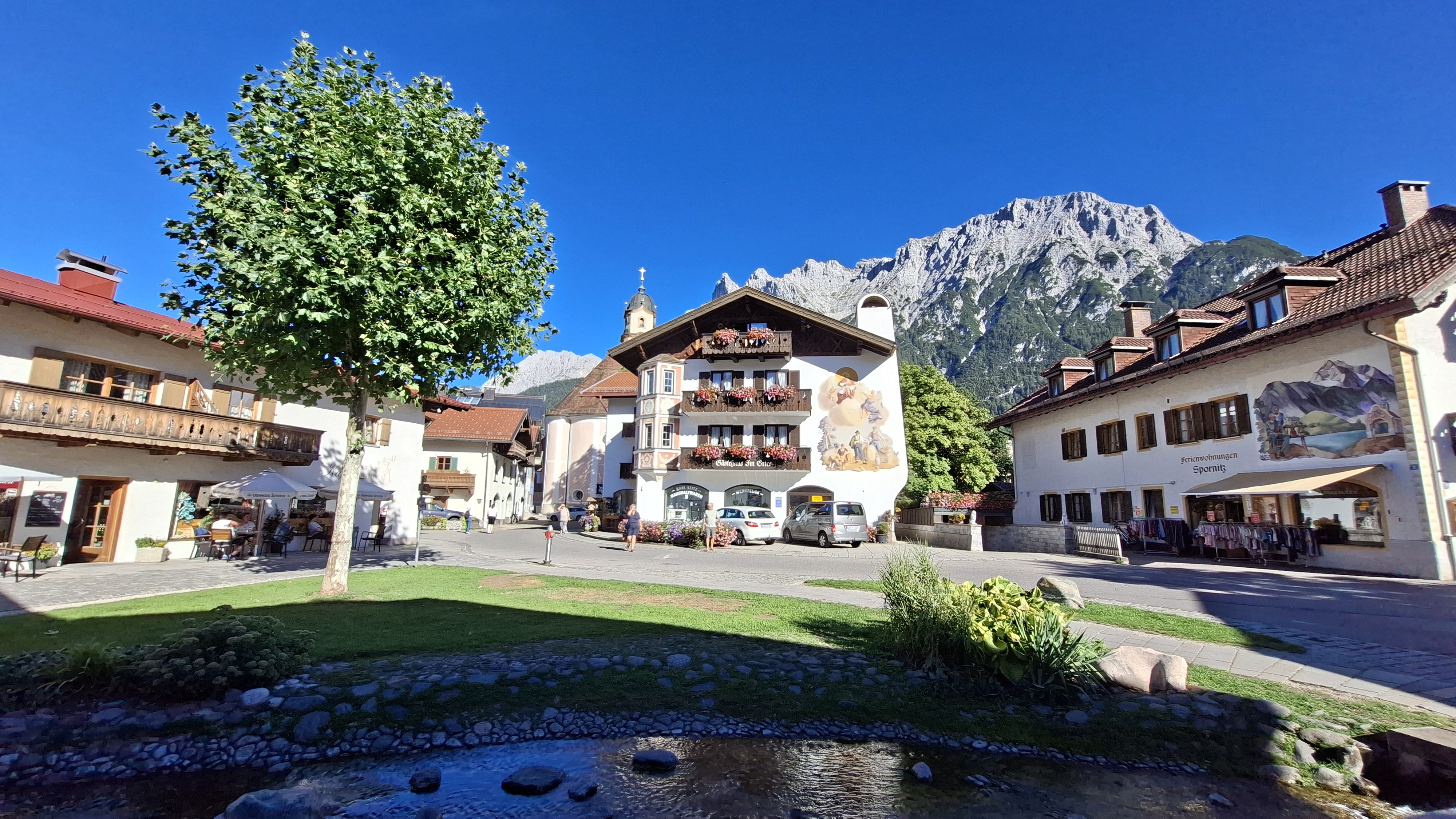 Painted facade of the Gästehaus Im Gries on Mittenwald's main square, with the parish church tower, a stream in the foreground, and the Karwendel peaks behind.