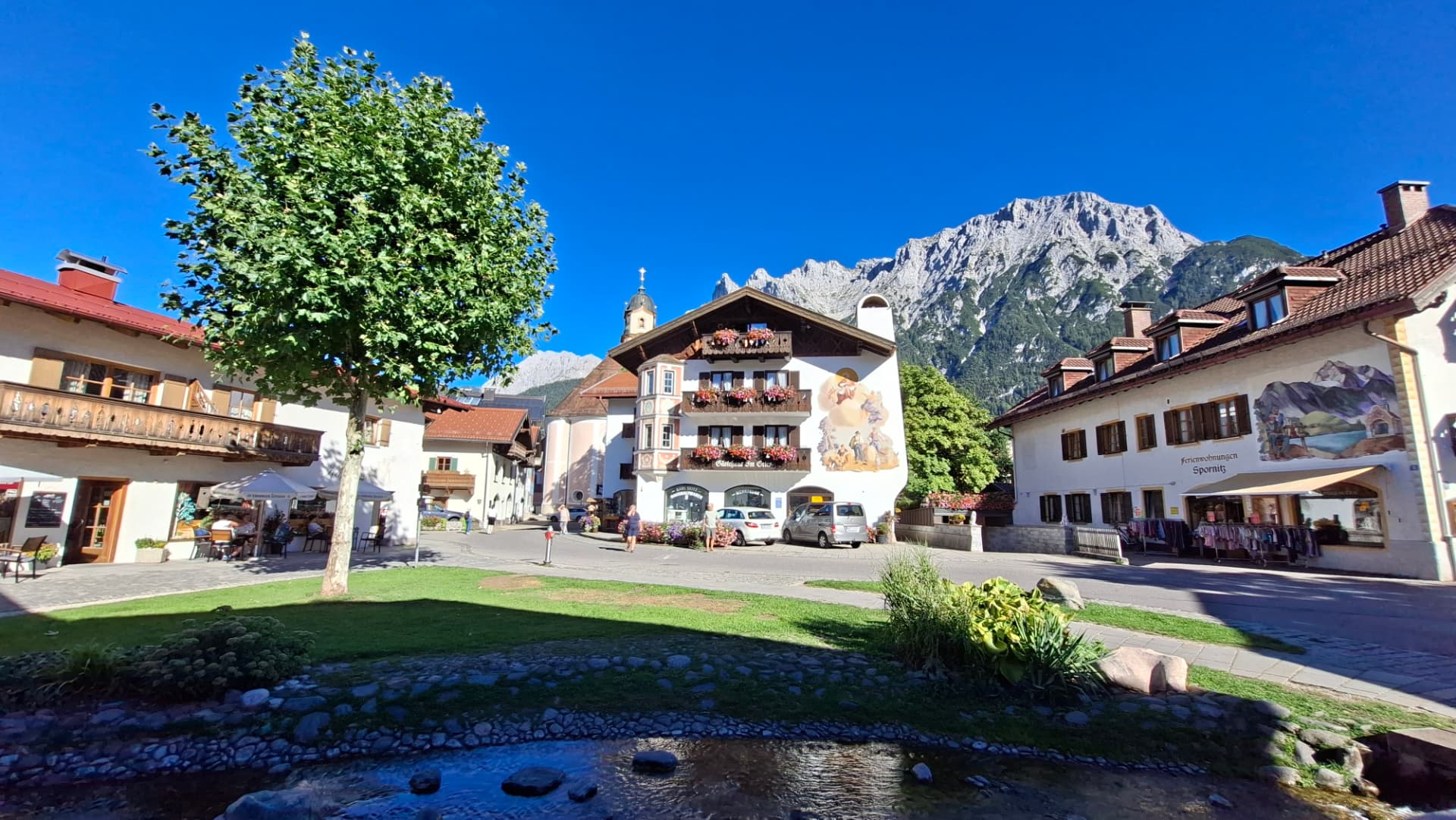 Painted facade of the Gästehaus Im Gries on Mittenwald's main square, with the parish church tower, a stream in the foreground, and the Karwendel peaks behind.